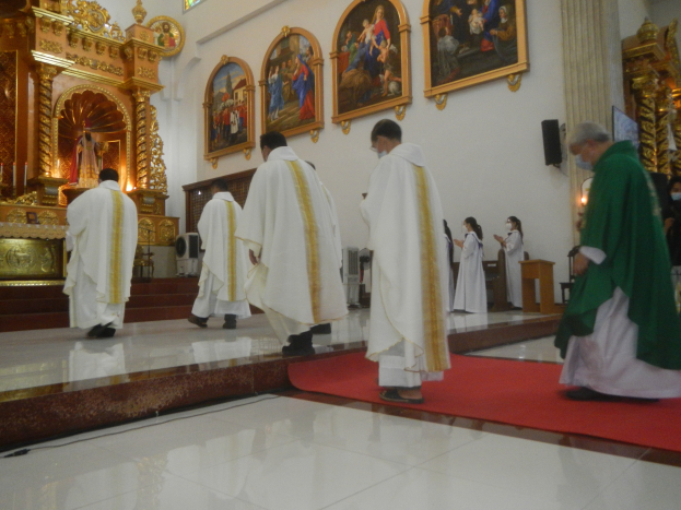 Eine Gruppe von Priestern geht einen roten Teppich in einer Kirche entlang, mit Menschen, die auf dem Boden und an den Wänden mit Fotorahmen im Hintergrund stehen.