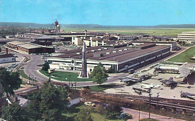 Altes Schwarz-Weiß-Foto eines Flughafens mit geparkten Flugzeugen, Gebäuden, Bäumen, Straßen, Fahrzeugen, Strommasten, Gras, Bahnschienen, Türmen, Hügeln und einem klaren Himmel, mit Text am unteren Rand.
