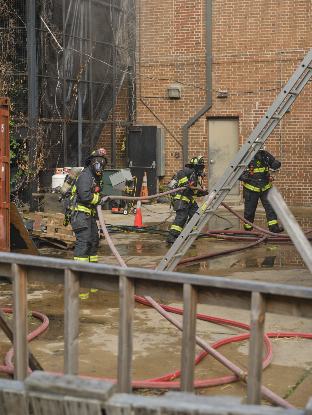 Feuerwehrleute in Helmen arbeiten daran, ein Gebäude Feuer zu löschen, während sie Schläuche halten, mit einem Metallzaun, Rohren, einem Behälter und einem Verkehrskegel in der Nähe.