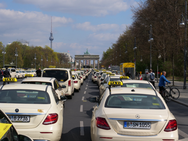Eine lange Reihe von Taxis parkt entlang einer belebten Straße in Berlin, Deutschland, mit Fahrradfahrern und Fußgängern auf dem Gehweg, flankiert von Bäumen und Gebäuden und einem Bogen und einem Turm im Hintergrund unter einem bewölkten Himmel.