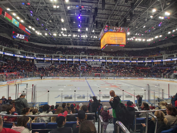 Eine gro├če Menschenmenge in einer Hockeyarena, mit einigen sitzend und anderen stehend, die ein Spiel unter hellen Lichtern und von der Decke h├Ąngenden Bannern beobachten, mit einem Zaun um die Eisbahn herum.