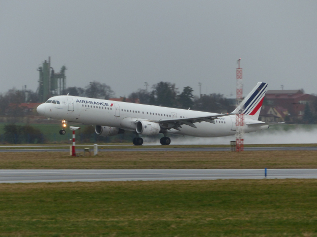 Air France Airbus A320-200 beim Start vom Paris Charles de Gaulle Airport, umgeben von grünem Gras und Bäumen, mit Gebäuden und Türmen im Hintergrund und einem klaren blauen Himmel.