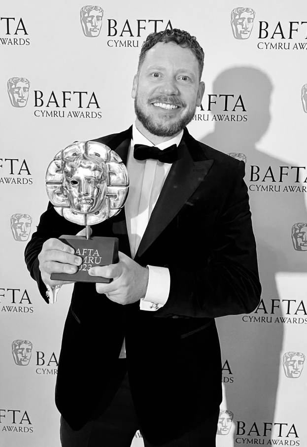 Ein Mann in einem Smoking, der eine BAFTA-Trophäe hält und lächelt, mit einem Hintergrund, auf dem 'BAFTA Awards 2018' steht.