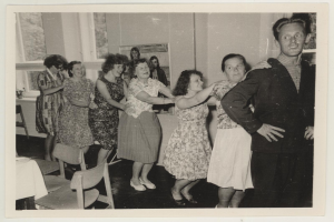 Eine Gruppe von Menschen, die in einem Raum im 1950er-Stil tanzen, mit Stühlen, einem Tisch, einem Fotoраhmen an der Wand und Fenstern im Hintergrund.