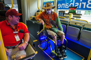 Zwei Männer im Rollstuhl auf einem Bus, einer trägt ein rotes T-Shirt und eine Kappe, der andere ein orangefarbenes Hemd und einen braunen Hut, beide tragen Masken und Ausweise, mit gelben Stangen und Glasfenstern, die Bäume, Gebäude und den Himmel zeigen.