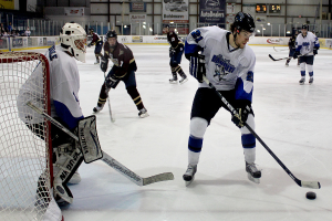 Eine Gruppe junger Männer, die Eishockey auf einem Eisplatz spielen, tragen Helme und halten Hockey-Schläger, mit einem Torpfosten links und Bannern im Hintergrund.