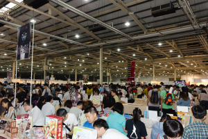 Eine große Gruppe von Menschen umringt Tische, die mit Büchern, Kisten und anderen Gegenständen gefüllt sind, auf einer Buchmesse.