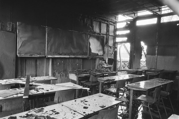 Schwarzes und weißes Foto eines Klassenzimmers mit verkohlten Wänden, zerbrochenen Fenstern und verstreuten Tischen und Stühlen.