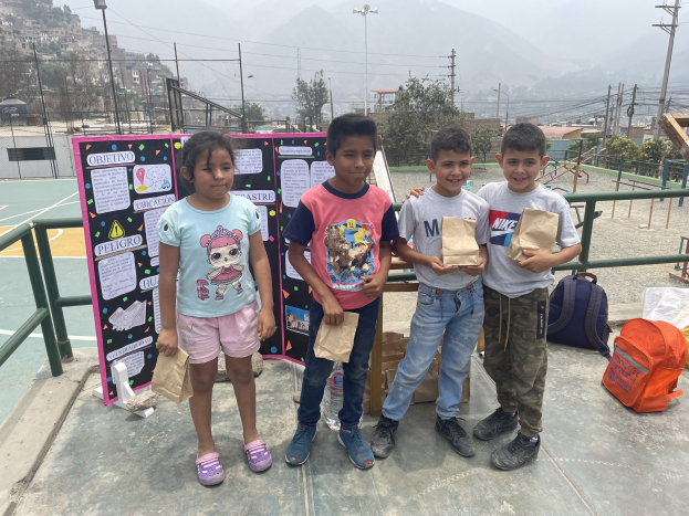 Gruppe von Kindern auf einem Basketballfeld, die Papiertüten halten, mit Tüten und Gegenständen rechts und einer Tafel mit Text links, vor einer Außenkulisse mit Zaun, Pfosten, Gebäuden.