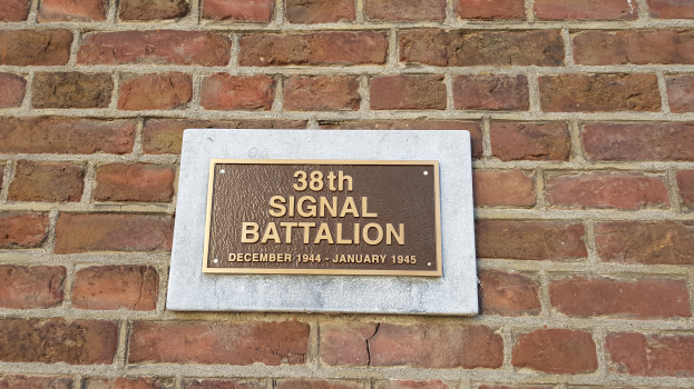 Eine Ziegelwand mit einer rechteckigen Plakette, auf der in fetter, weißer, zentrierter Schrift "38. Signal Battalion" steht.