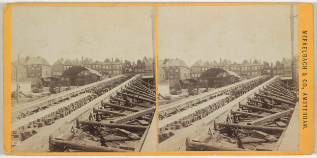 Ein altes Schwarz-Weiß-Foto einer Bahnschiene mit Gebäuden im Hintergrund, Text auf der rechten Seite, der 'Merkelbach & Co. Amsterdam' lautet, und verstreute hölzerne Gegenstände im Vordergrund.