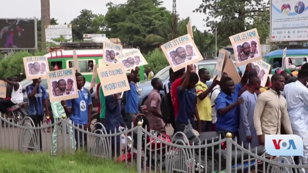 Eine Gruppe von Menschen mit Schildern, wahrscheinlich protestierend, steht vor einem Zaun mit Fahrzeugen, Büschen, Pfosten, Gittern und einem Bildschirm im Hintergrund, unter einem sichtbaren Himmel mit Gras am unteren Bildrand.