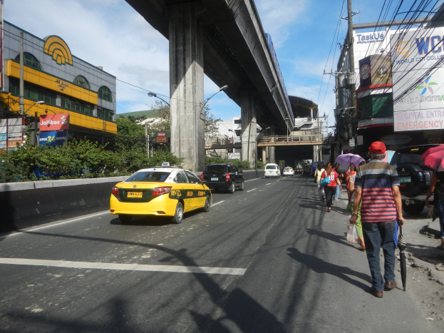 Ein gelbes Taxi fährt eine Stadtstraße mit hohen Gebäuden, Fußgängern mit Regenschirmen, einer Brücke im Hintergrund, bewölktem Himmel und Oberleitungen.