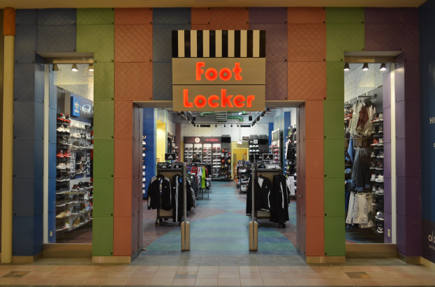 Innenansicht eines Foot Locker-Schuhgeschäfts mit Kleidung an Bügeln und Gestellen unter beleuchteter Beleuchtung, wobei der Boden sichtbar ist.