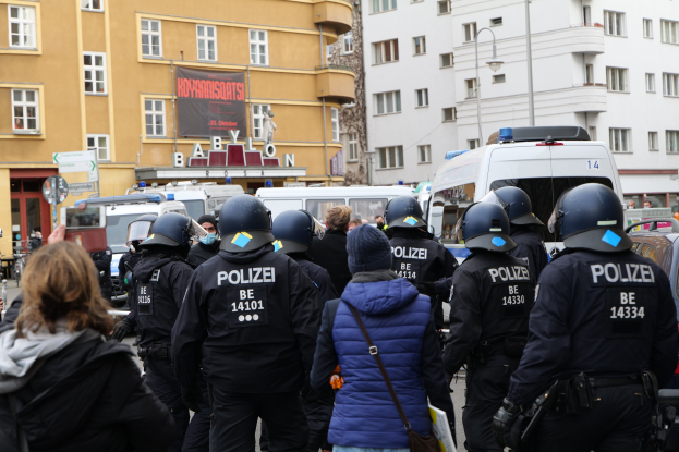 Polizeibeamte in Uniform vor einer Menge von Helm- und Jackenträgern während einer Demonstration in Berlin, mit Fahrzeugen, Gebäuden, Laternenmasten und einem Banner im Hintergrund.