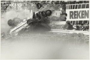 Ein Schwarz-Weiß-Foto eines demolierten Rennwagens, umgeben von Reifen, mit einer Gruppe von Menschen rechts und einer Tafel im Hintergrund.