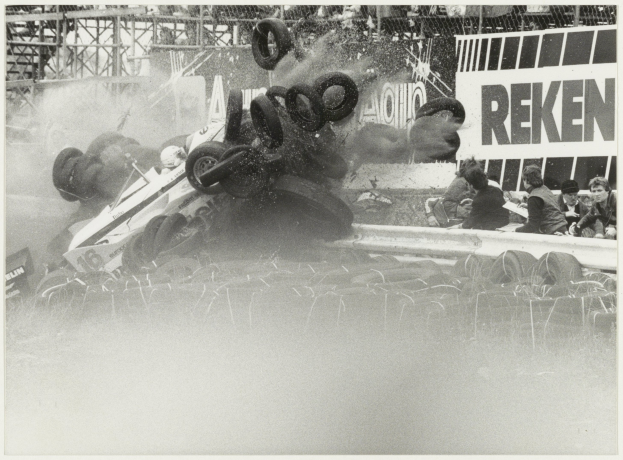 Ein Schwarz-Weiß-Foto eines demolierten Rennwagens, umgeben von Reifen, mit einer Gruppe von Menschen rechts und einer Tafel im Hintergrund.