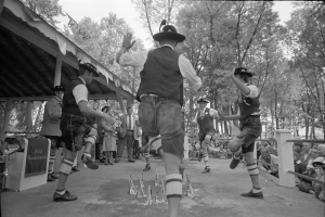 Eine Gruppe von Männern in traditioneller bayrischer Tracht tanzt auf einer Bühne vor einem gemischten Publikum aus sitzenden und stehenden Menschen, mit Bäumen und einer Hütte im Hintergrund, dargestellt in Schwarz-Weiß.