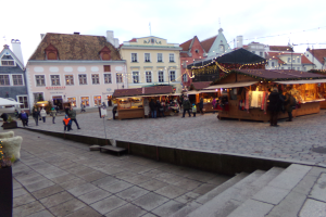 Ein lebendiger Weihnachtsmarkt in Tallinn, Estland, mit Menschen um geschmückte Stände, festlicher Beleuchtung und Gebäuden mit Fenstern im Hintergrund, unter einem bewölkten Himmel, mit Stufen, die zum Markt führen, und Töpfen mit Pflanzen, die herumstehen.