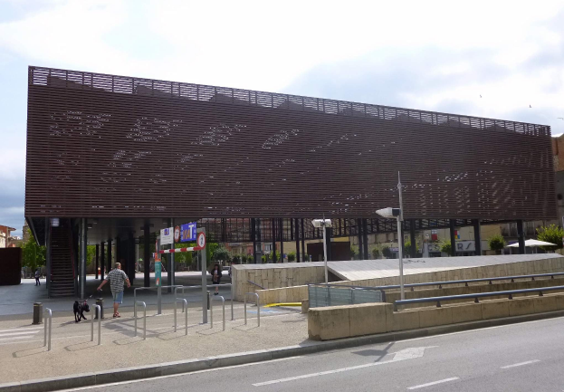Großes modernes Gebäude mit zahlreichen Fenstern, das als neuer Eingang zum Stadtzentrum dient, flankiert von Straßeninfrastruktur, Fahrzeugen, Fußgängern und Bäumen unter einem bewölkten Himmel.