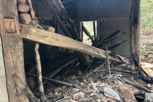 Ein altes Gebäude umgeben von Trümmern wie Holzstöcke, Steine und trockene Blätter, mit Bäumen im Hintergrund, scheint im Abrissprozess zu sein.