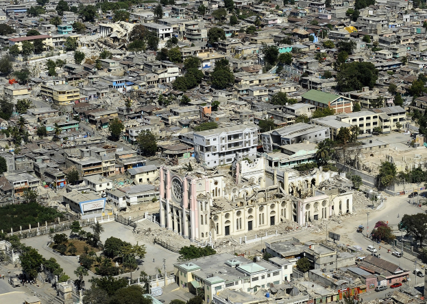 Luftaufnahme einer Stadt mit einer zerstörten Kirche im Zentrum, umgeben von Gebäuden, Bäumen, Straßen, Fahrzeugen und Trümmern nach einem Erdbeben.