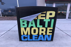 Schwarzer Müllcontainer mit 'Keep Baltimore More Clean'-Text auf einem Gehweg vor einem Gebäude mit Glastüren.