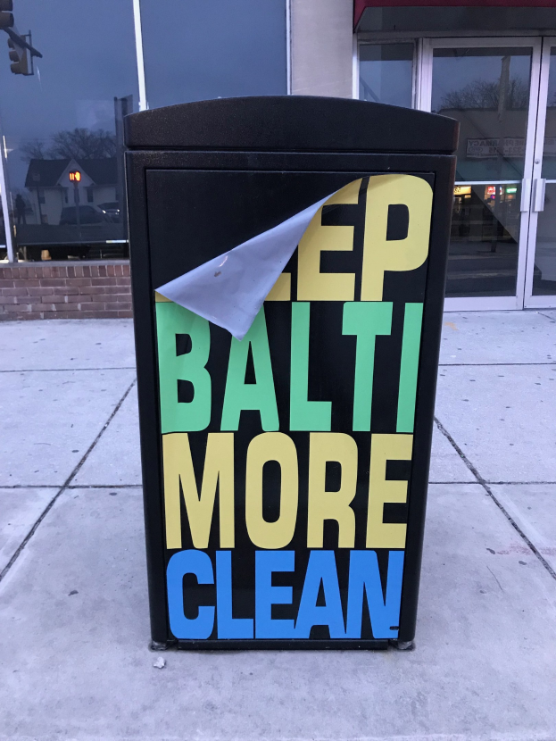 Schwarzer Müllcontainer mit 'Keep Baltimore More Clean'-Text auf einem Gehweg vor einem Gebäude mit Glastüren.