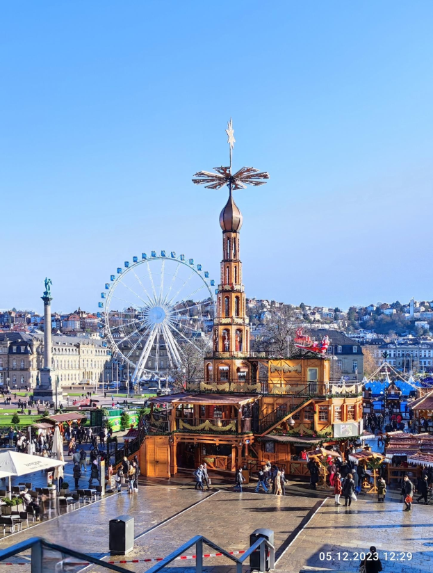 Ein belebter Stadtplatz mit einem zentralen Riesenrad, umgeben von Gebäuden, Bäumen und Menschen unter einem klaren blauen Himmel, mit Marktzelten und Geländern.