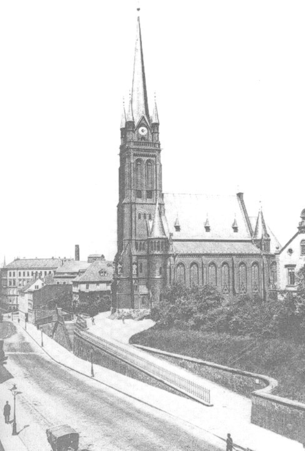 Schwarzes und weißes Foto einer Stadtstraße mit der St. Peter Kirche in Wiesbaden, Deutschland im Hintergrund, gesäumt von Gebäuden, Bäumen, Fahrzeugen und einigen Fußgängern.