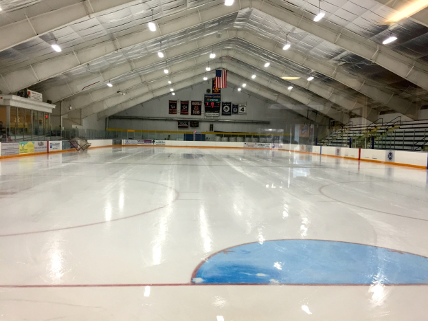 Große Indoor-Eisfläche mit Deckenbeleuchtung, Werbetafeln, Treppen mit Geländern, einer Wand im Hintergrund und einigen Menschen, was auf eine Sportanlage hinweist, die likely für Hockey verwendet wird.