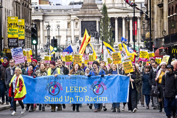 Menschen marschieren mit einer "Steh Auf gegen Rassismus"-Tafel die Straße entlang, mit Gebäuden, Bäumen, einer Statue und einem bewölkten Himmel im Hintergrund.