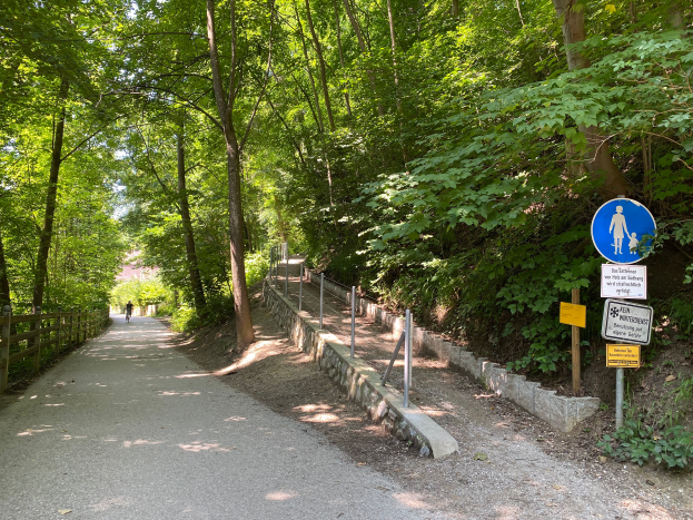 Eine Person fährt mit dem Fahrrad auf einem asphaltierten Weg durch den Wald, mit Bäumen und einem hölzernen Zaun auf der linken Seite und Schildern mit Text und Bildern auf der rechten Seite.