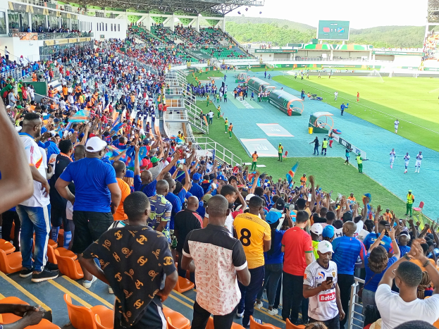 Eine große Menschenmenge versammelt sich vor einem eingezäunten Fußballfeld mit Hügeln und einem klaren blauen Himmel im Hintergrund und wartet gespannt auf den Beginn eines Spiels während der Afrikameisterschaft 2019.