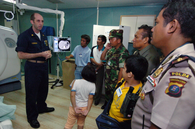 Eine Gruppe von Menschen, die um ein Krankenhauszimmer herumstehen, mit einem Mann, der in der Mitte ein Mikrofon hält, einem Bett mit einer Decke darauf links, verschiedenen medizinischen Geräten, einem Röntgengerät, einer Tafel mit Text, einer Tür, einer Wand und einer Decke mit Deckenleuchten im Hintergrund.