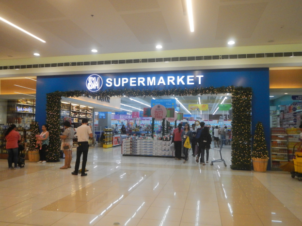 Eine Gruppe von Menschen steht vor einem Supermarkt, einige halten Taschen, mit einem Einkaufswagen, Topfpflanze, Regalauslagen, Beschilderung und Deckenbeleuchtung im Bild.