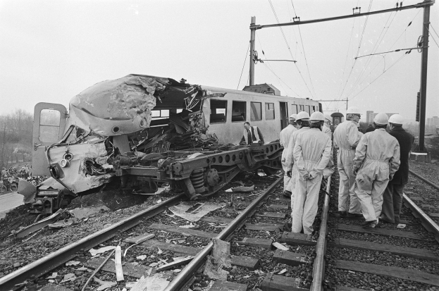 Schwarzes und weißes Bild einer Zugkollision auf den Gleisen mit Einsatzkräften in Helmen und einem Hintergrund aus Strommasten, Bäumen, Gebäuden und einem klaren Himmel.