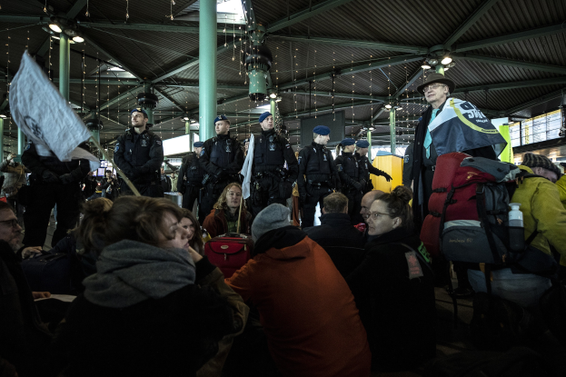Eine Gruppe von Menschen vor einer Menge an einem Bahnhof, einige tragen Mützen und tragen Taschen, mit Säulen und Deckenlampen im Hintergrund; die Menge scheint zu protestieren, hält Schilder und Transparente.
