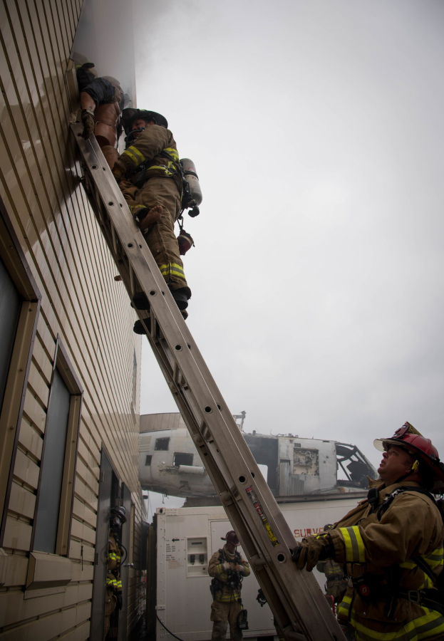 Feuerwehrleute mit Helmen, die ein Gebäude mit einer Leiter erklimmen, mit einem Fahrzeug und dem Himmel im Hintergrund.