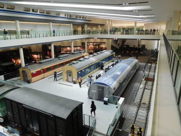 Ein großzügiger Museumsausstellungsraum mit verschiedenen Zügen in unterschiedlichen Größen und Farben, bei dem Besucher vom Boden, Geländern und Säulen aus beobachten, unter Deckenbeleuchtung.