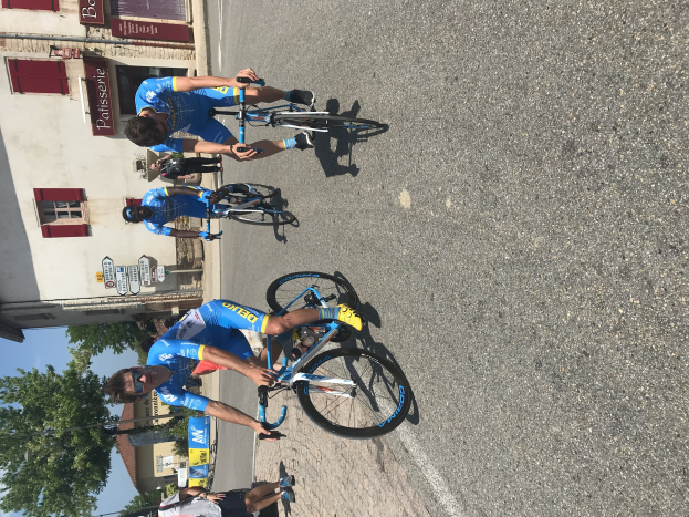 Eine Gruppe von Radfahrern fährt eine Straße entlang an einem Gebäude mit vernagelten Fenstern und Text vorbei, Bäumen und einem klaren Himmel im Hintergrund, mit Zuschauern auf der Straße darunter.
