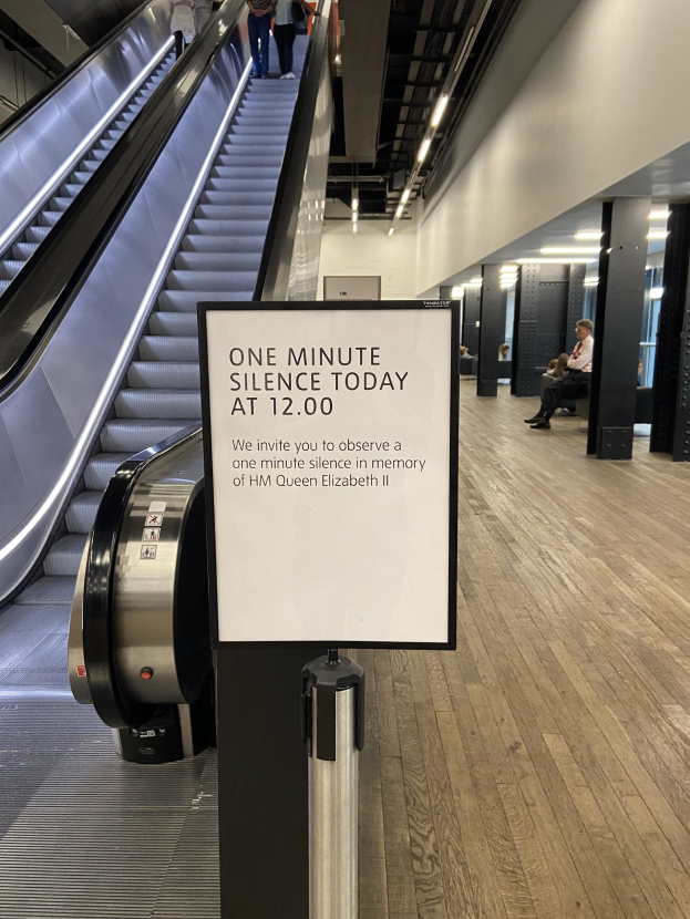 Eine Rolltreppe in einem Flughafen mit einem Schild, auf dem "Eine Minute Stille heute" steht, einige Menschen darauf und Lampen an der Decke im Hintergrund.