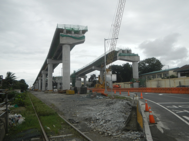 Baustelle mit einer Brücke im Hintergrund, Straße mit Absperrgittern markiert, Bahnschiene links, verstreute Steine und Gras, Bäume und Gebäude säumen die Straße und ein bewölkter Himmel.
