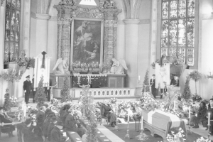 Schwarz-weißes Foto einer Trauerfeier in einer Kirche mit Sitzplätzen und Stehplätzen, Blumensträußen, Kerzen, einem Sarg im Vordergrund und einem gerahmten Foto und Skulpturen im Hintergrund.