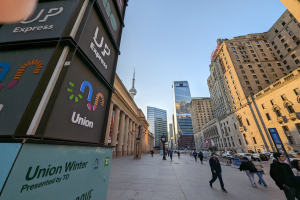 Eine belebte Straße mit Fussgängern, hohen Geb"uden, Fahrzeugen und Texttafeln auf der linken Seite, unter einem sichtbaren Himmel.