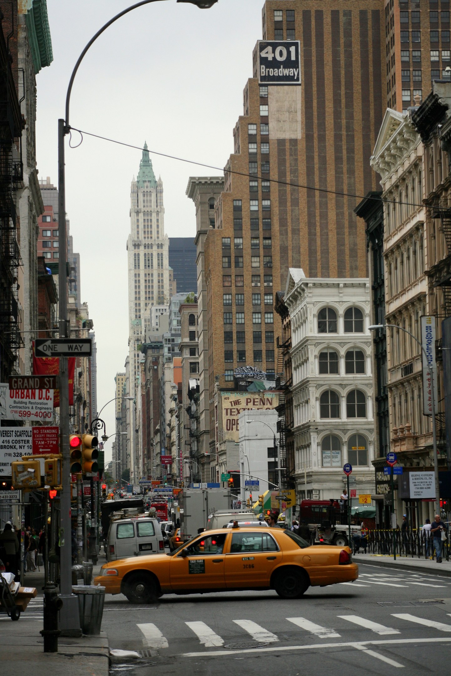Ein gelbes Taxi fährt auf einer von Hochhäusern gesäumten Stadtstraße, Fußgänger auf dem Gehweg, Ampeln und Texttafeln unter einem sichtbaren Himmel.
