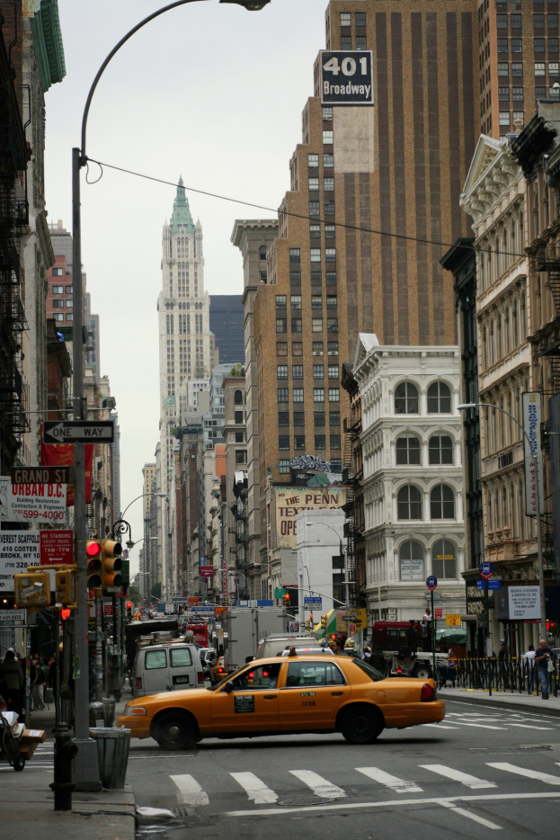 Ein gelbes Taxi fährt auf einer von Hochhäusern gesäumten Stadtstraße, Fußgänger auf dem Gehweg, Ampeln und Texttafeln unter einem sichtbaren Himmel.