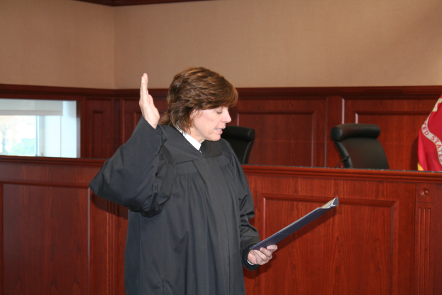 Eine Frau in einer schwarzen Robe steht an einem Richtertisch und hält eine Akte in der Hand, mit einem Mikrofon und einer Flagge auf dem Tisch und Stühlen und einem Fenster dahinter.