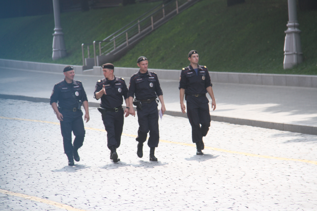 Drei Polizisten in Uniform, die eine Straße entlanggehen, mit Laternen, Geländern, Treppen und Gras im Hintergrund.