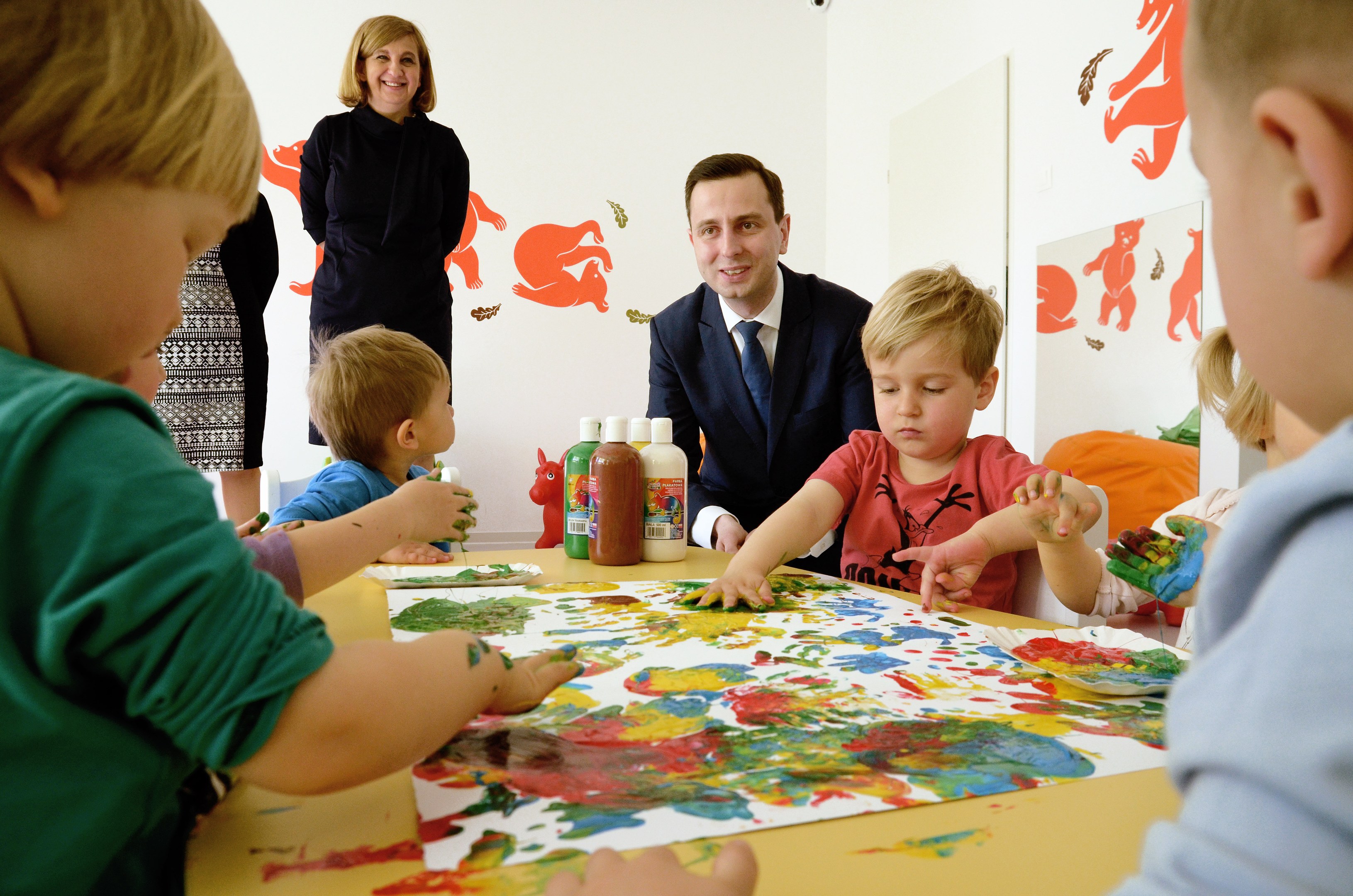 Gruppe von Kindern bei einer Malaktivität an einem Tisch mit Farbe an den Händen, Papieren und Flaschen, während eine Frau im Hintergrund mit Gemälden an der Wand lächelt.